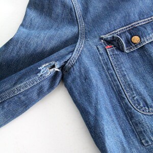 K&ouml;nnte beinhalten: Eine blaue Jeansjacke mit einem einzigen Knopfverschluss und einer aufgesetzten Tasche auf der linken Seite. Die Jacke hat einen abgenutzten Look mit einem kleinen Loch im linken &Auml;rmel.