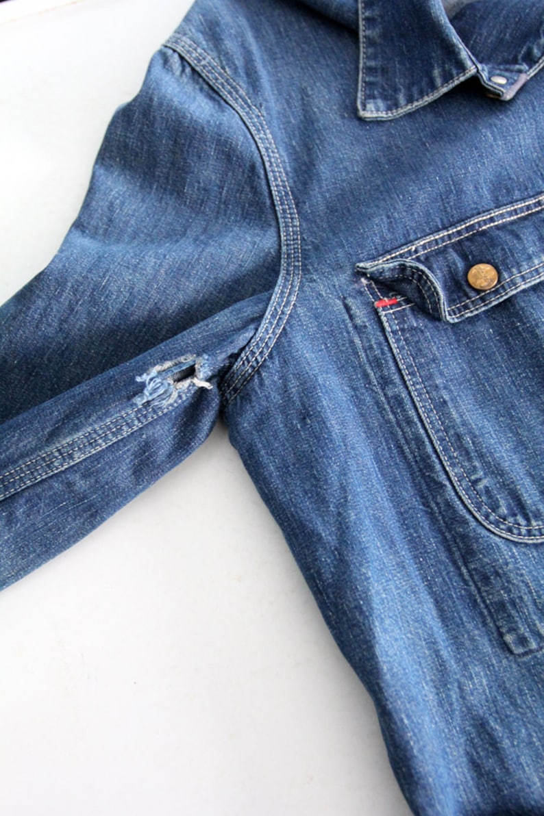 K&ouml;nnte beinhalten: Eine blaue Jeansjacke mit einem einzigen Knopfverschluss und einer aufgesetzten Tasche auf der linken Seite. Die Jacke hat einen abgenutzten Look mit einem kleinen Loch im linken &Auml;rmel.
