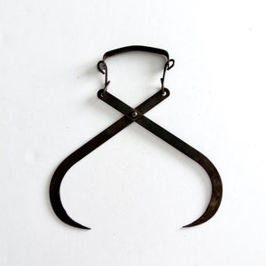 Puede incluir: Una pinza para hielo vintage de metal oscuro con un asa curvada y puntas afiladas. Las pinzas están cruzadas en el centro y parecen ser de hierro o acero. El fondo es blanco liso.
