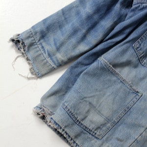 K&ouml;nnte beinhalten: Nahaufnahme einer blauen Jeansjacke mit ausgefranstem Saum und einer aufgesetzten Tasche. Die Jacke ist abgetragen und hat einen ausgebleichten Look.