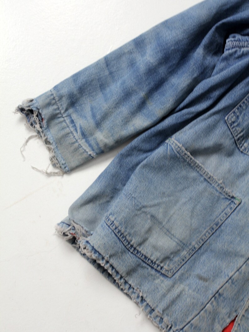 K&ouml;nnte beinhalten: Nahaufnahme einer blauen Jeansjacke mit ausgefranstem Saum und einer aufgesetzten Tasche. Die Jacke ist abgetragen und hat einen ausgebleichten Look.
