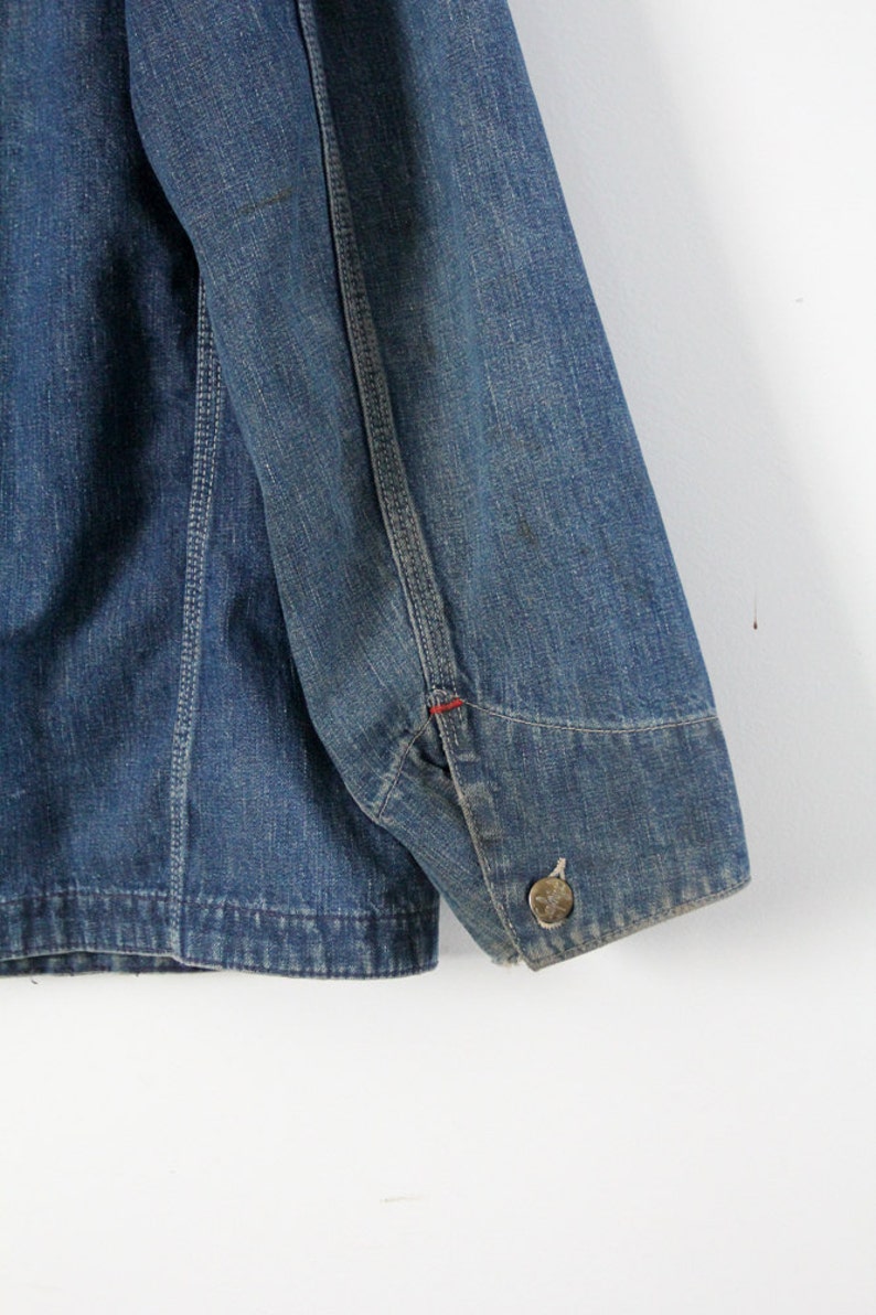 K&ouml;nnte beinhalten: Eine blaue Jeansjacke mit einem roten Stichdetail am &Auml;rmel. Die Jacke hat einen Knopfverschluss und einen einzigen Knopf am &Auml;rmel.