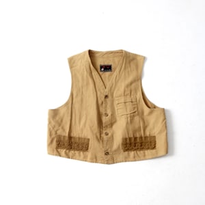 Puede incluir: Un chaleco de algodón de color beige con cierre de botones y dos bolsillos. El chaleco tiene un ribete marrón alrededor de los bolsillos.