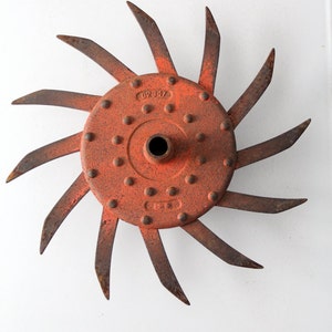 Könnte beinhalten: Ein rostiges, orangefarbenes Metall-Fräsmesser mit einem zentralen Loch und mehreren gebogenen Klingen, die nach außen strahlen. Die Klinge hat eine kreisförmige Mitte mit kleinen Nieten und der Zahl "02847". Die Klingen sind spitz und weisen Gebrauchsspuren auf.