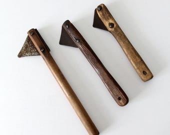May include: Three small vintage hatchets with wooden handles and metal blades. The hatchets have varying shades of brown and are arranged diagonally against a white background. The blades are triangular and attached to the handles with rivets.