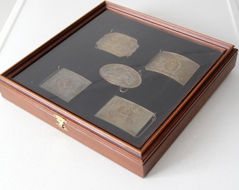 Puede incluir: Una vitrina de madera con tapa de cristal, que muestra cinco hebillas de cinturón de metal vintage. Las hebillas son de varias formas y tamaños, con diseños y textos intrincados. La caja tiene un interior oscuro y un cierre de latón.