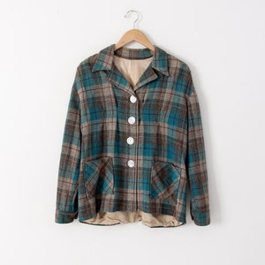 May include: A vintage plaid jacket in teal, brown, and gray, with large white buttons and two front pockets. The jacket is hanging on a wooden hanger against a white background. The jacket has a collared neckline.