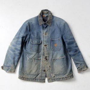 Op de afbeelding: Vintage blauwe denim werkjas met een versleten uiterlijk. Voorzien van een knoopsluiting, twee borstzakken en twee onderste zakken. De kraag heeft een donkerdere blauwtint. De jas vertoont tekenen van slijtage, met beschadigde plekken op de mouwen en zakken.