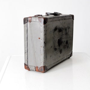 May include: Vintage gray suitcase with metal corner protectors and rivets. The suitcase has a black handle and a latch closure. The exterior shows signs of wear, adding to its antique charm. A classic travel accessory.