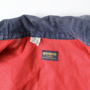 K&ouml;nnte beinhalten: Ein rotes gestepptes Futter mit einem dunkelblauen Cordkragen. Ein wei&szlig;es Etikett mit dem Text "OshKosh B'Gosh" und "Sanforized" ist in das Futter eingen&auml;ht.