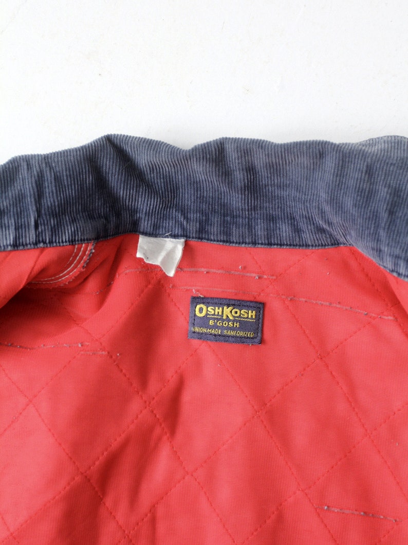 K&ouml;nnte beinhalten: Ein rotes gestepptes Futter mit einem dunkelblauen Cordkragen. Ein wei&szlig;es Etikett mit dem Text "OshKosh B'Gosh" und "Sanforized" ist in das Futter eingen&auml;ht.