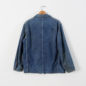 K&ouml;nnte beinhalten: Eine blaue Jeansjacke mit Knopfleiste und langen &Auml;rmeln. Die Jacke h&auml;ngt an einem Holzb&uuml;gel vor einem wei&szlig;en Hintergrund.