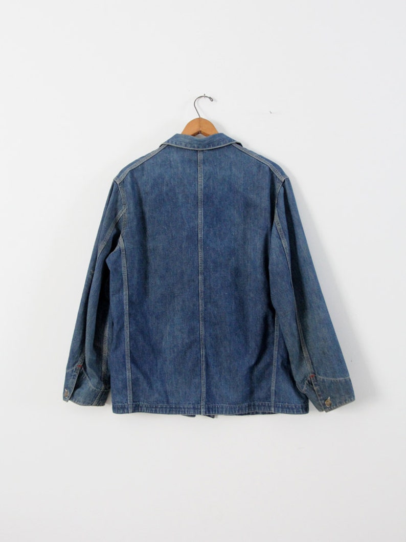 K&ouml;nnte beinhalten: Eine blaue Jeansjacke mit Knopfleiste und langen &Auml;rmeln. Die Jacke h&auml;ngt an einem Holzb&uuml;gel vor einem wei&szlig;en Hintergrund.