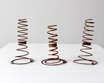 May include: Three vintage metal springs with a spiral design. The springs are a rusty brown color and stand upright on a white surface. Each spring has a circular base.