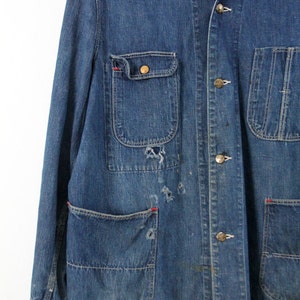 K&ouml;nnte beinhalten: Eine blaue Jeans-Arbeitsjacke mit mehreren Taschen und Messingkn&ouml;pfen. Die Jacke hat einen abgenutzten Look mit sichtbarer Ausbleichung und N&auml;hten.