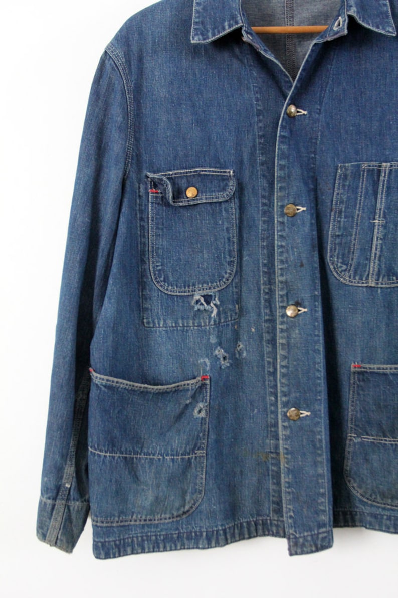 K&ouml;nnte beinhalten: Eine blaue Jeans-Arbeitsjacke mit mehreren Taschen und Messingkn&ouml;pfen. Die Jacke hat einen abgenutzten Look mit sichtbarer Ausbleichung und N&auml;hten.
