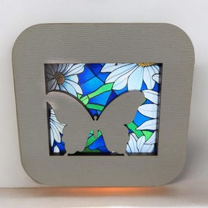 May include: A gray wooden frame with a cutout butterfly shape. The frame contains a stained glass image of a butterfly with blue, green, and white flowers.