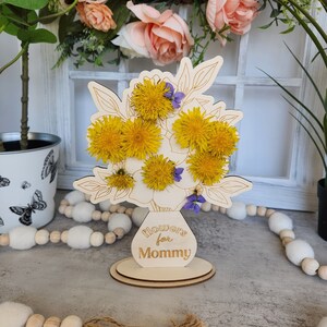 May include: Wooden floral arrangement with yellow dandelions and purple flowers. The arrangement is in a vase-shaped cutout with the text "Flowers for Mommy". The base is round. The background includes a window and decorative elements.