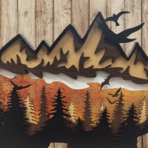 May include: A wooden wall art piece depicting a mountain range with a sunset behind it. The mountains are brown and the sunset is orange and yellow. There are trees in the foreground.