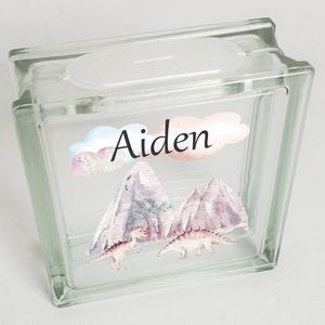 Puede incluir: Huchas de vidrio con un dise&ntilde;o de acuarela de monta&ntilde;as, dinosaurios y nubes. El nombre "Aiden" est&aacute; escrito en la parte delantera.