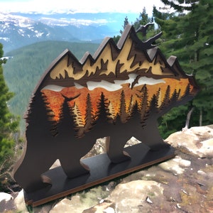 May include: A wooden bear silhouette with a layered mountain scene inside. The bear is standing on a wooden base. The mountain scene is painted in shades of orange, yellow, and brown. The background is a blurry image of a mountain range.