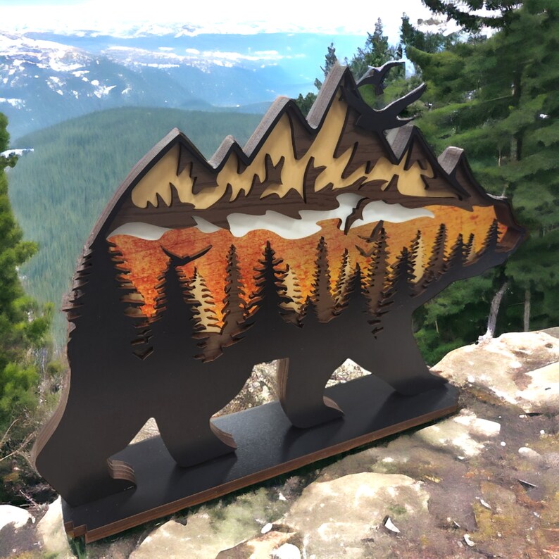 May include: A wooden bear silhouette with a layered mountain scene inside. The bear is standing on a wooden base. The mountain scene is painted in shades of orange, yellow, and brown. The background is a blurry image of a mountain range.