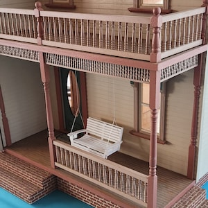 May include: A white wooden porch swing hangs from the ceiling of a dollhouse porch. The porch has a wooden railing and a brick floor. The swing is made of white wood with a slatted back and seat.