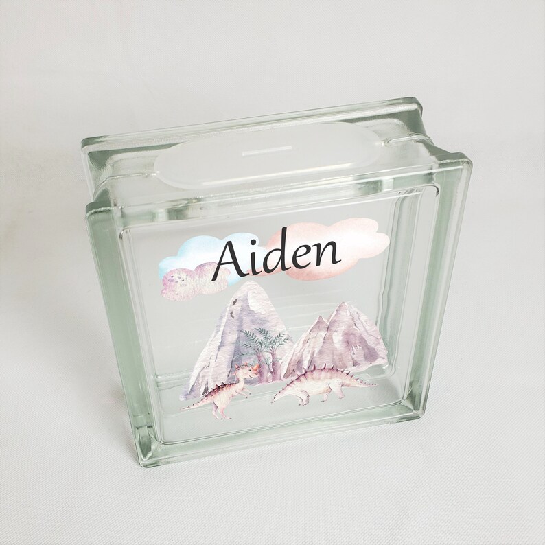 Puede incluir: Una alcanc&iacute;a de vidrio transparente con una ranura en la parte superior. La parte delantera de la alcanc&iacute;a tiene una pegatina colorida con una escena de monta&ntilde;a, dos dinosaurios y el nombre "Aiden".