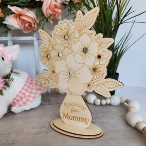 May include: A wooden floral arrangement with the text "flowers for Mommy" engraved on the vase. The flowers have mirrored centers and are set on a wooden base. The design is light brown.