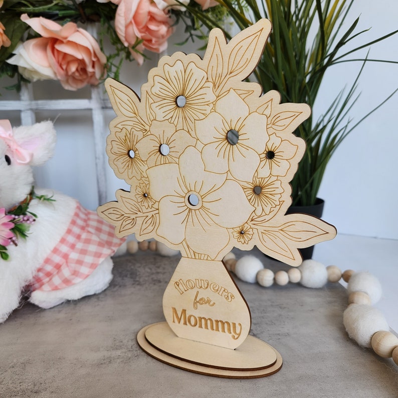 May include: A wooden floral arrangement with the text "flowers for Mommy" engraved on the vase. The flowers have mirrored centers and are set on a wooden base. The design is light brown.