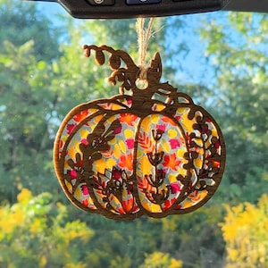 May include: A wooden pumpkin-shaped car ornament with a fall-themed design. The pumpkin is painted with a variety of colors, including red, orange, yellow, and green. The ornament is perfect for adding a touch of fall decor to your car.