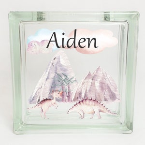 Puede incluir: Un bloque de vidrio con un dise&ntilde;o de acuarela de monta&ntilde;as, &aacute;rboles y dinosaurios. El nombre "Aiden" est&aacute; escrito en letras cursivas negras en el bloque.