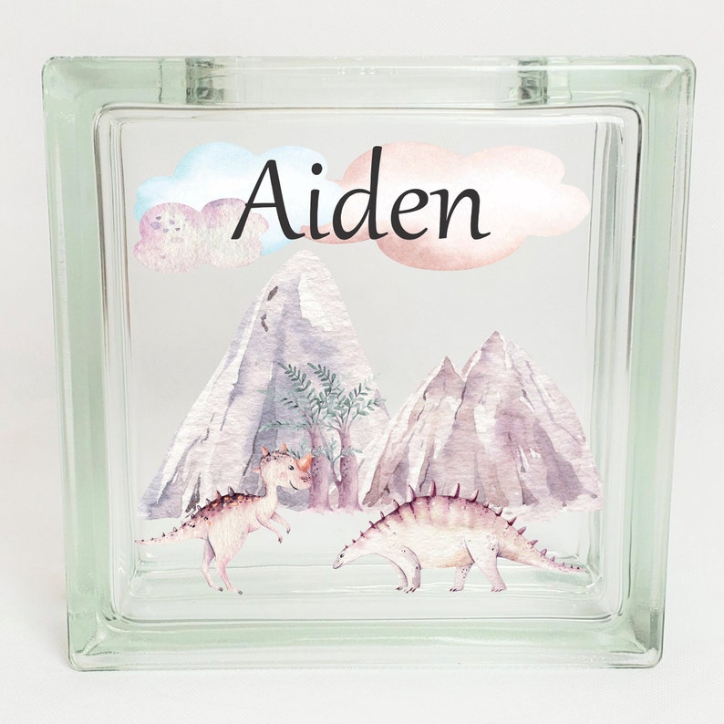 Puede incluir: Un bloque de vidrio con un dise&ntilde;o de acuarela de monta&ntilde;as, &aacute;rboles y dinosaurios. El nombre "Aiden" est&aacute; escrito en letras cursivas negras en el bloque.