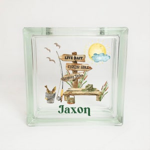 May include: A glass block with a personalized design featuring a fishing scene. The design includes a wooden sign with the words "Live Bait", "Fishin' Hole", and "Weighing Station", a fishing rod, a bucket of fish, a bench, a hat, and a sun. The name "Jaxon" is written in green at the bottom of the block.