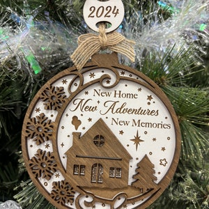 May include: A wooden Christmas ornament with a white background and brown accents. The ornament features a house, a tree, snowflakes, and stars. The text "New Home New Adventures New Memories" is written on the ornament. The year "2024" is written on a separate tag.