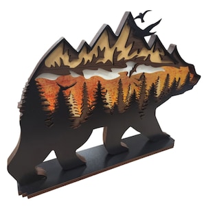 May include: Wooden bear silhouette with a layered mountain and forest scene inside. The scene features a sunset with orange, yellow, and red hues, and silhouettes of trees. There are also several birds flying in the sky.
