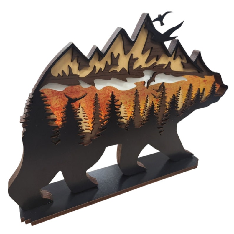 May include: Wooden bear silhouette with a layered mountain and forest scene inside. The scene features a sunset with orange, yellow, and red hues, and silhouettes of trees. There are also several birds flying in the sky.