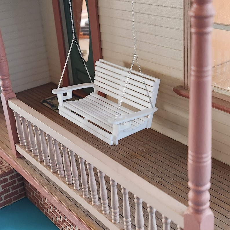 May include: White wooden porch swing hanging from a chain on a wooden porch with a white railing. The swing has a slatted back and seat.