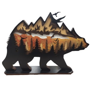 May include: A wooden bear silhouette with a layered design of mountains, trees, and a sunset. The bear is standing on a wooden base.