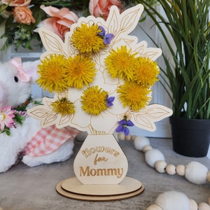 May include: Wooden floral arrangement with yellow dandelions and purple flowers. The arrangement is in a vase-shaped base with the words "flowers for Mommy" engraved on it. The base is round and made of wood.