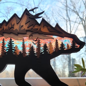 May include: A wooden silhouette of a bear with a mountain range and a forest scene inside. The bear is black and the mountain range is brown. The forest scene is a colorful abstract design with blue, green, and orange colors.