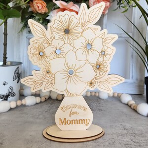 May include: A wooden floral bouquet decoration with engraved details and small mirrored accents. The vase has the words "flowers for Mommy" engraved on it. The bouquet is on a round wooden base.