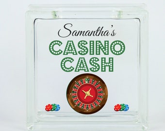 May include: A glass money jar with a green and white "Samantha's Casino Cash" design and a roulette wheel graphic. There are three red, blue, and green poker chips at the bottom of the jar.