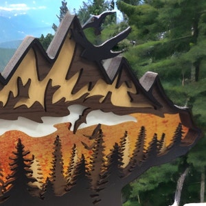 May include: A wooden wall art piece depicting a mountain range with a forest in the foreground. The mountains are brown and the forest is green. The sky is blue and there are clouds in the sky.