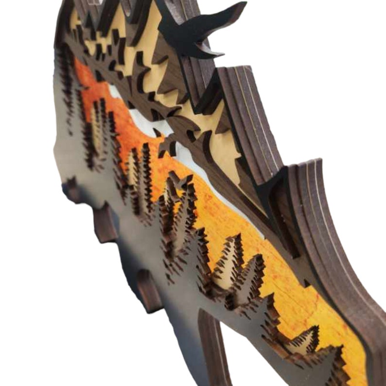 May include: A wooden wall art piece with a layered design of a mountain range with trees and a sunset sky. The piece is made of multiple layers of wood, creating a 3D effect.