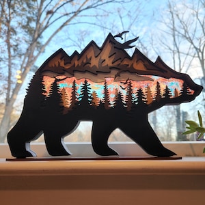 May include: A wooden bear silhouette with a layered mountain scene inside. The mountains are painted in shades of orange, red, and blue, with a hint of green for the trees. The bear is standing on a wooden base.
