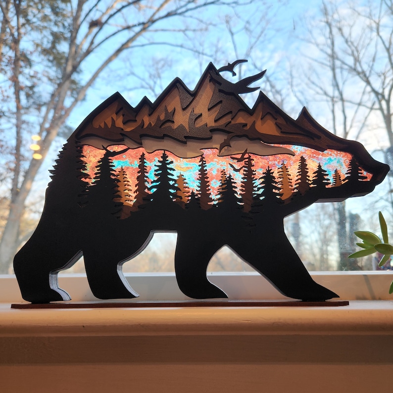 May include: A wooden bear silhouette with a layered mountain scene inside. The mountains are painted in shades of orange, red, and blue, with a hint of green for the trees. The bear is standing on a wooden base.