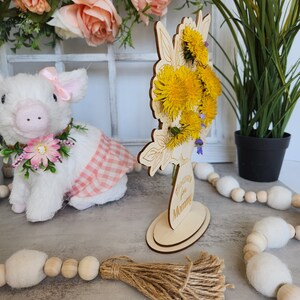 May include: A wooden flower vase with a floral design, holding yellow dandelions. The vase has the text "Wishing for Mommy". A stuffed pig wearing a pink bow and a pink and white checkered dress is in the background.