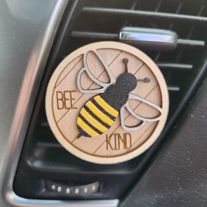 May include: A wooden car air freshener with a bee design. The bee is black and yellow with a striped abdomen. The words "bee kind" are written on the air freshener.