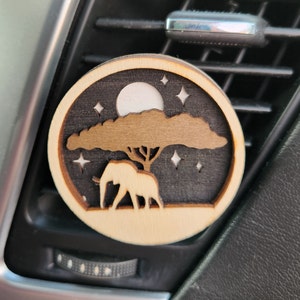 May include: A wooden car air freshener with a silhouette of an elephant standing under a tree with a full moon and stars in the background.